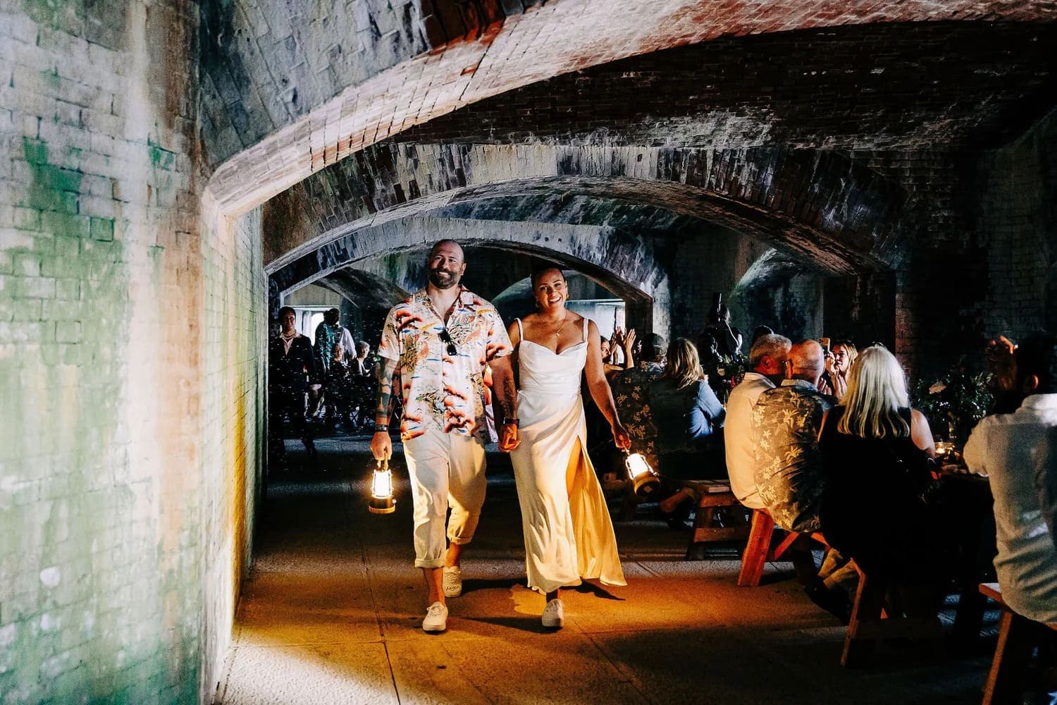 A couple joyfully walks hand-in-hand through a dimly lit archway, each holding a lantern.