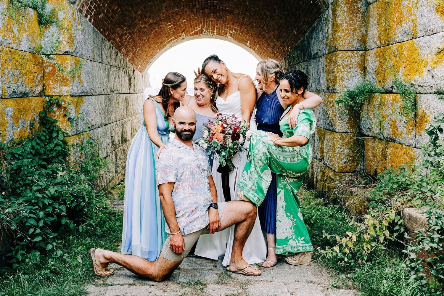 A joyful group of women in colorful dresses, along with a man, pose together under an arched stone pathway, celebrating a special occasion.