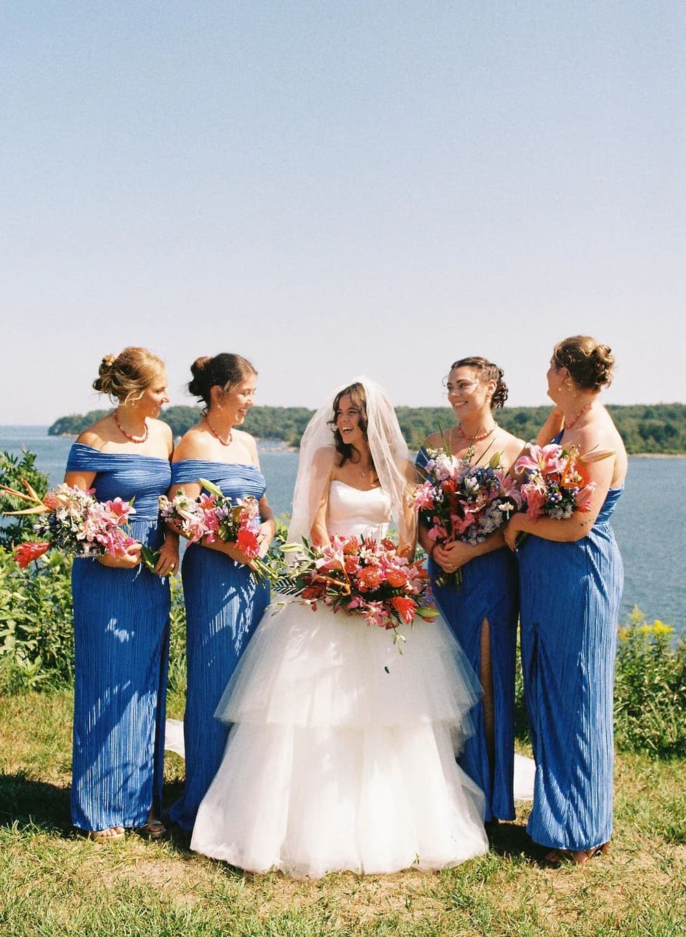 A bride in a white dress smiles joyfully surrounded by her bridesmaids in blue dresses, holding vibrant floral bouquets by a scenic waterfront.