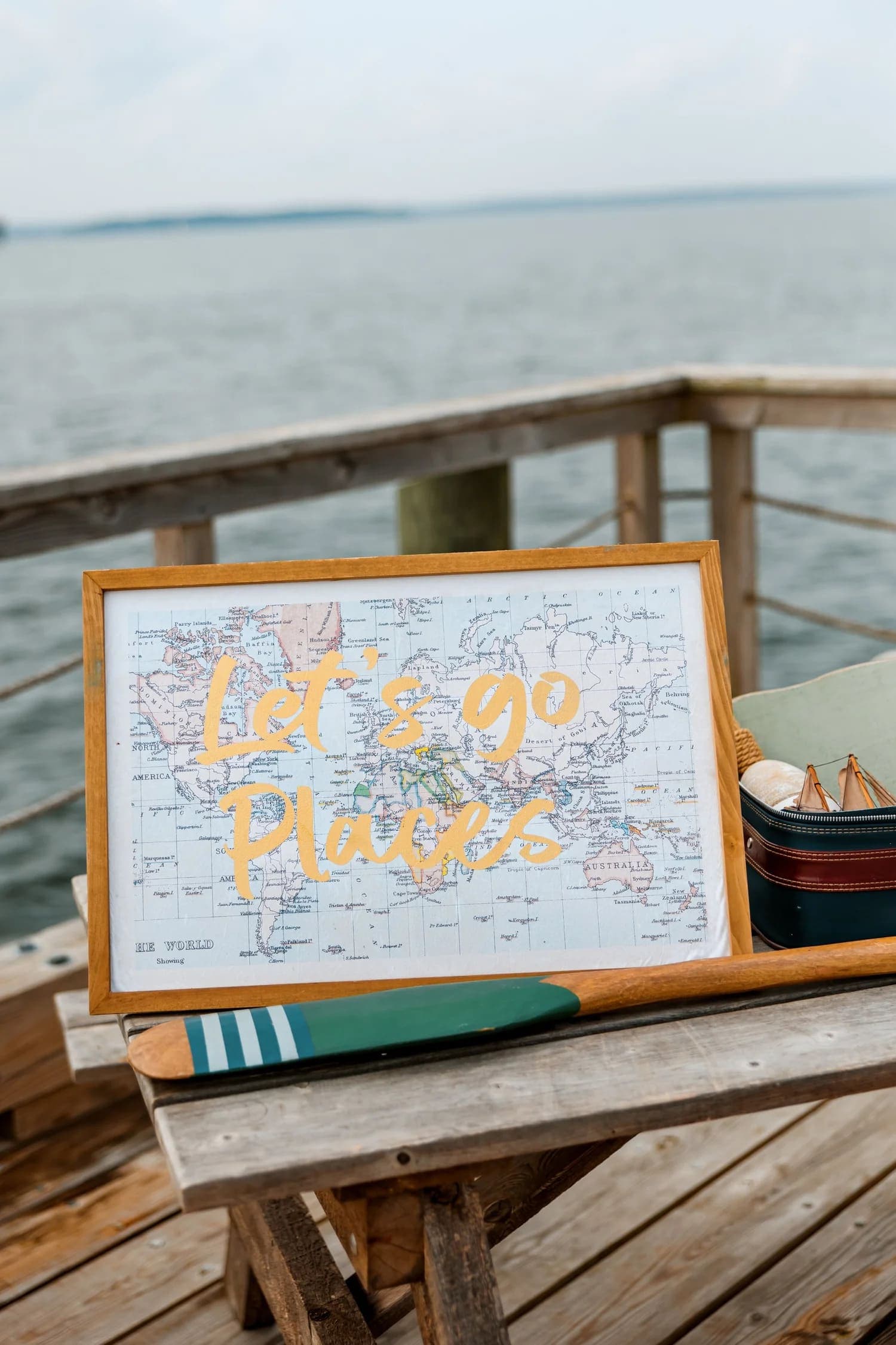 A framed map with the words "Let's go Places" displayed on a wooden table by the water.
