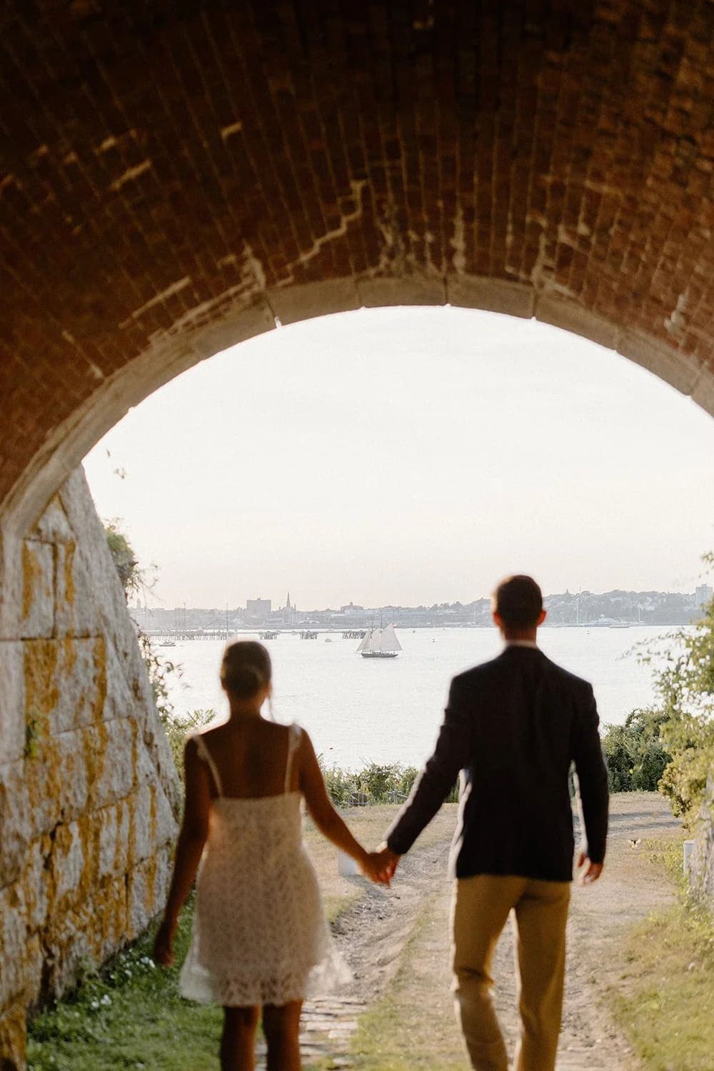 A couple walks hand in hand through an arched brick tunnel, leading to a scenic waterfront view.