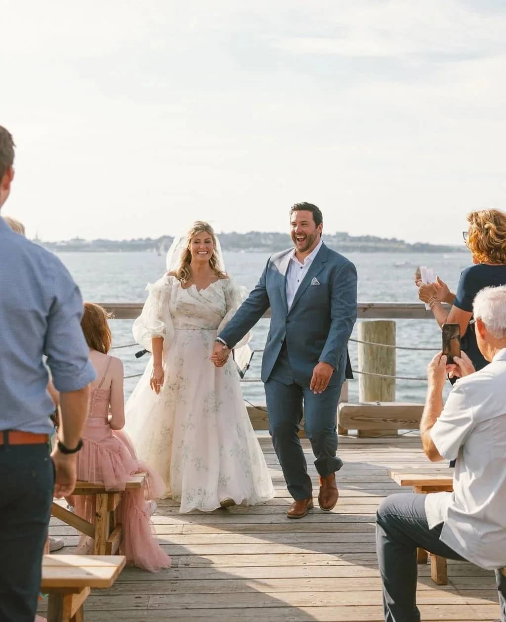 A bride and groom joyfully walk hand in hand down a waterfront wooden dock amid a cheering crowd.
