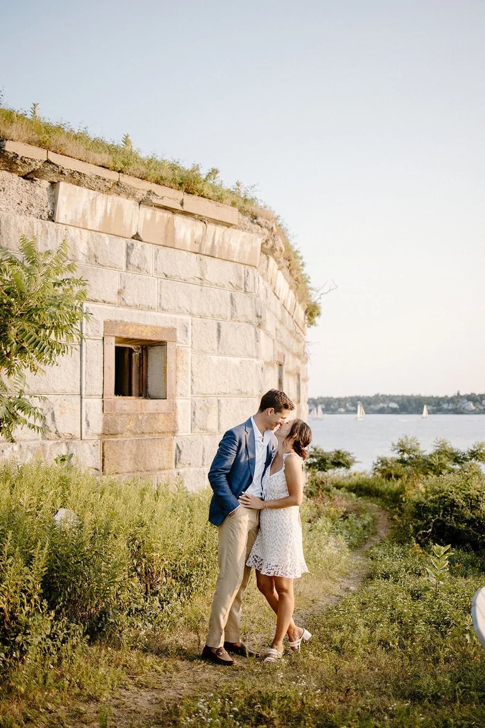 A couple embraces near a historic stone wall by the water during golden hour.