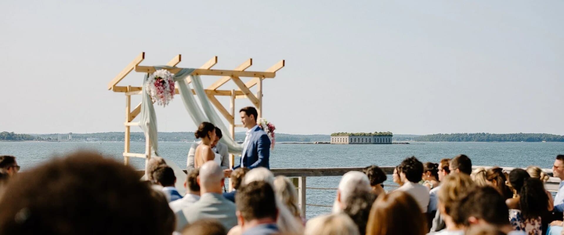 A couple exchanges vows under a wooden arch by the water, surrounded by wedding guests.