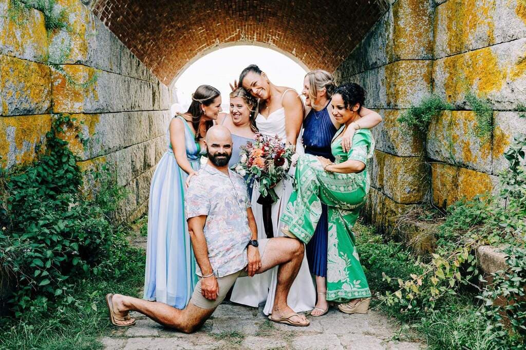 A group of six friends poses playfully in a lush, green outdoor setting, with one person in a wedding dress holding a bouquet.