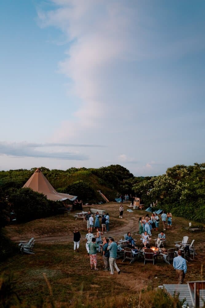 A lively outdoor gathering with people dressed in light clothing around a fire pit and a large tent under a clear sky.