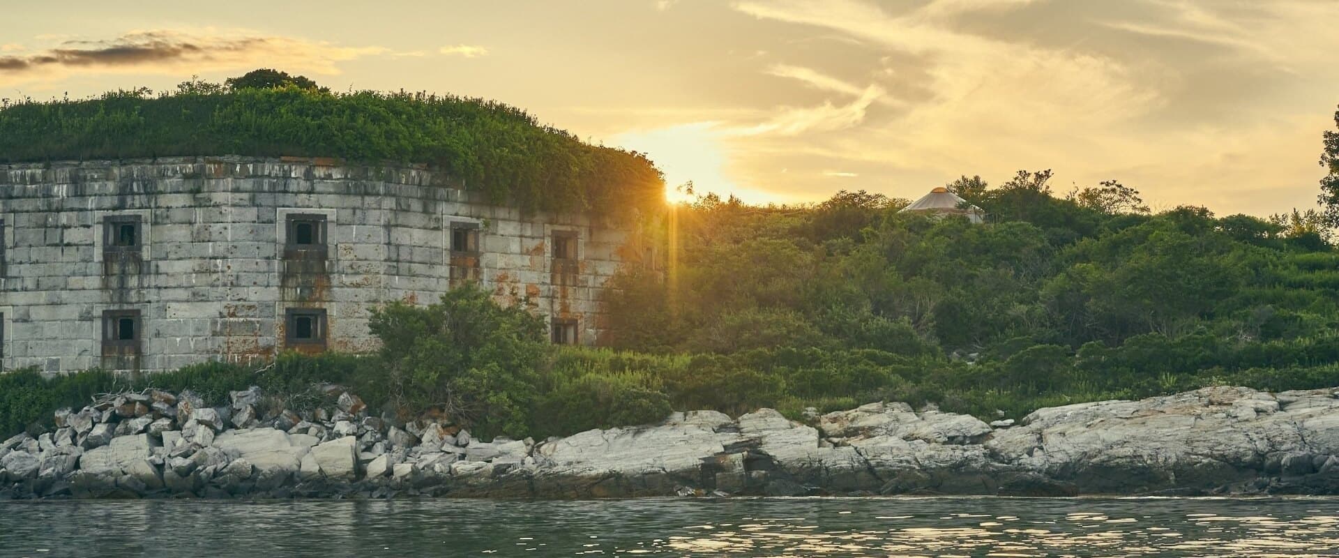 Sunset illuminates a weathered, overgrown fortress by the water's edge.