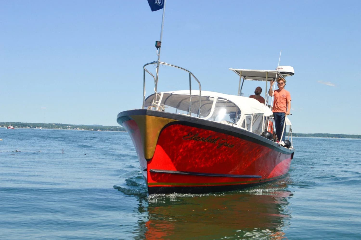 A red boat with two people aboard navigates calm water on a sunny day.