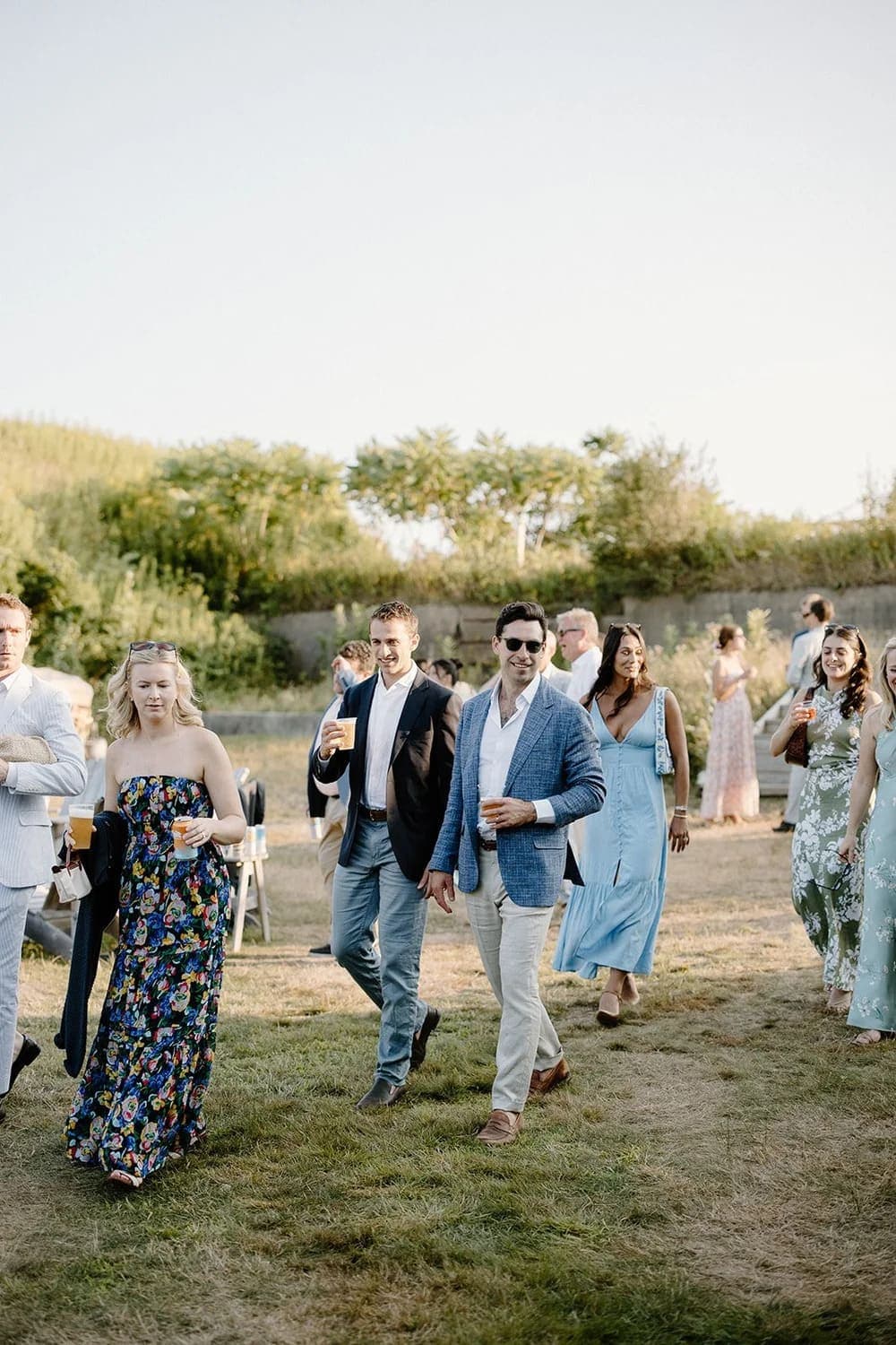 A group of people in formal attire enjoys drinks while walking through a green outdoor space.
