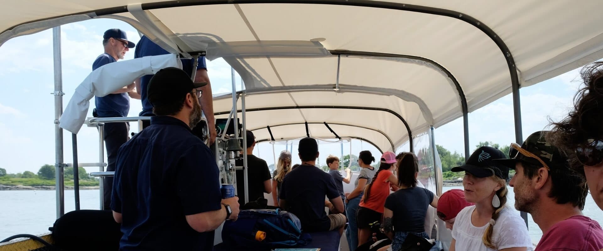 A group of people sitting on a covered boat, some listening to a speaker at the front.