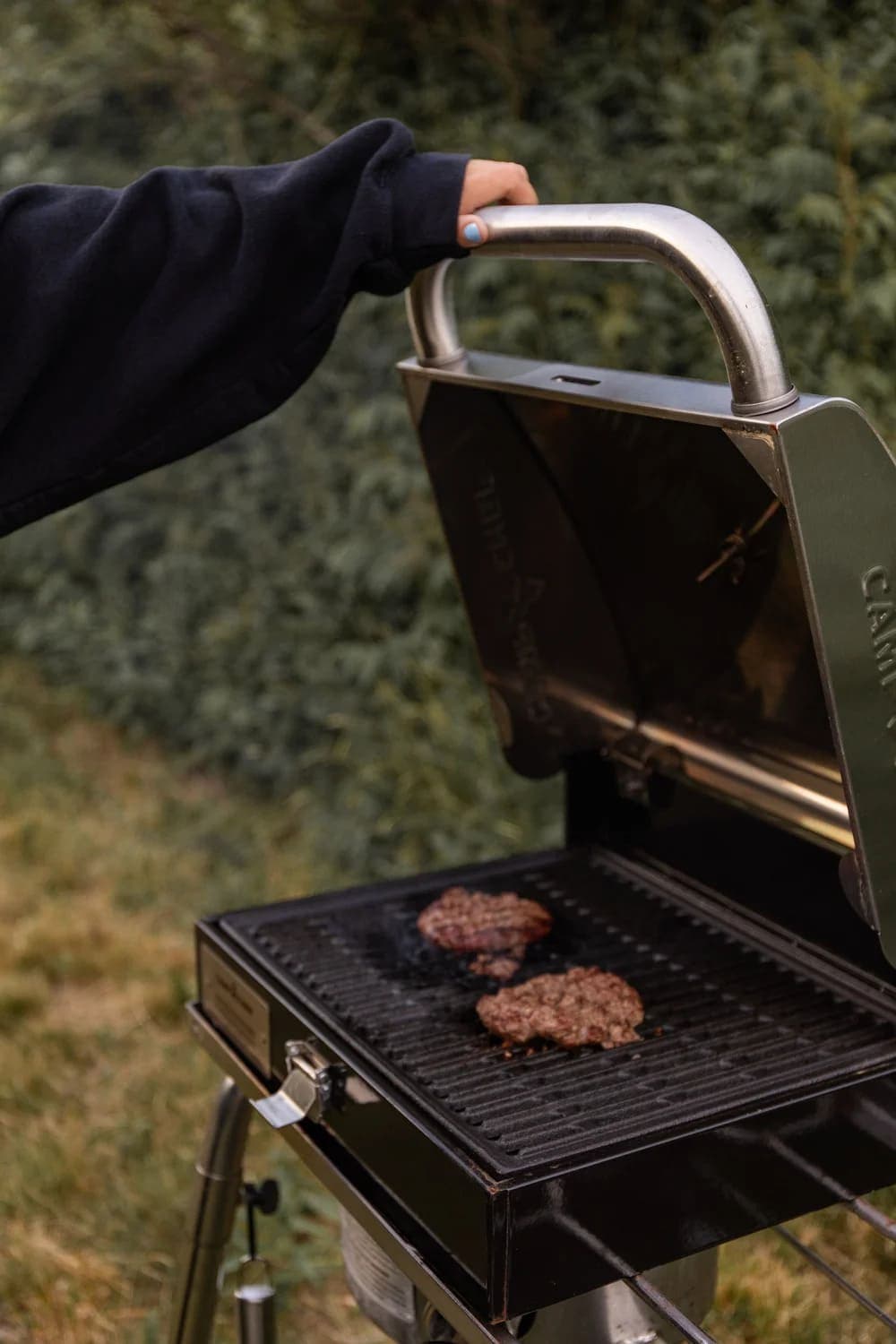 A hand in a black sweater raises the lid of a grill with burgers sizzling inside.