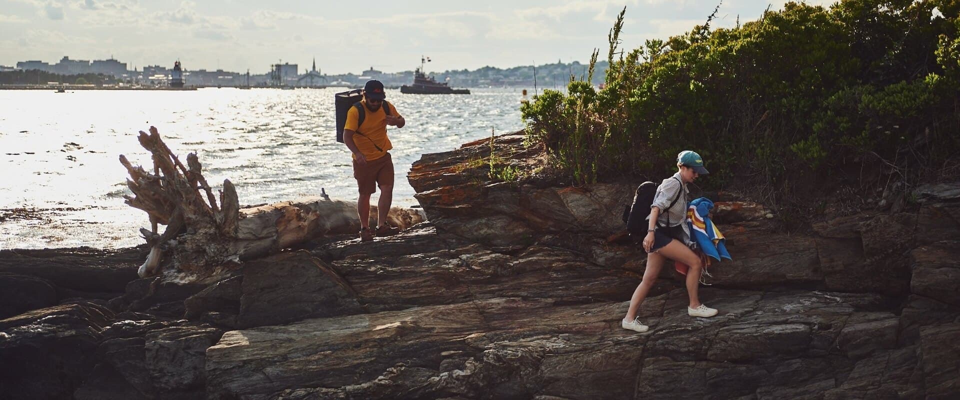 Two people navigate rocky terrain by the water under a cloudy sky.