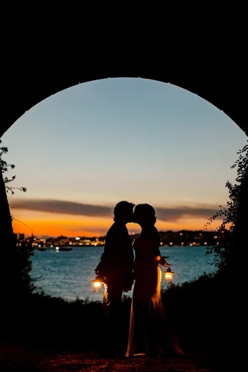 A couple silhouettes kiss while holding lanterns against a sunset backdrop.