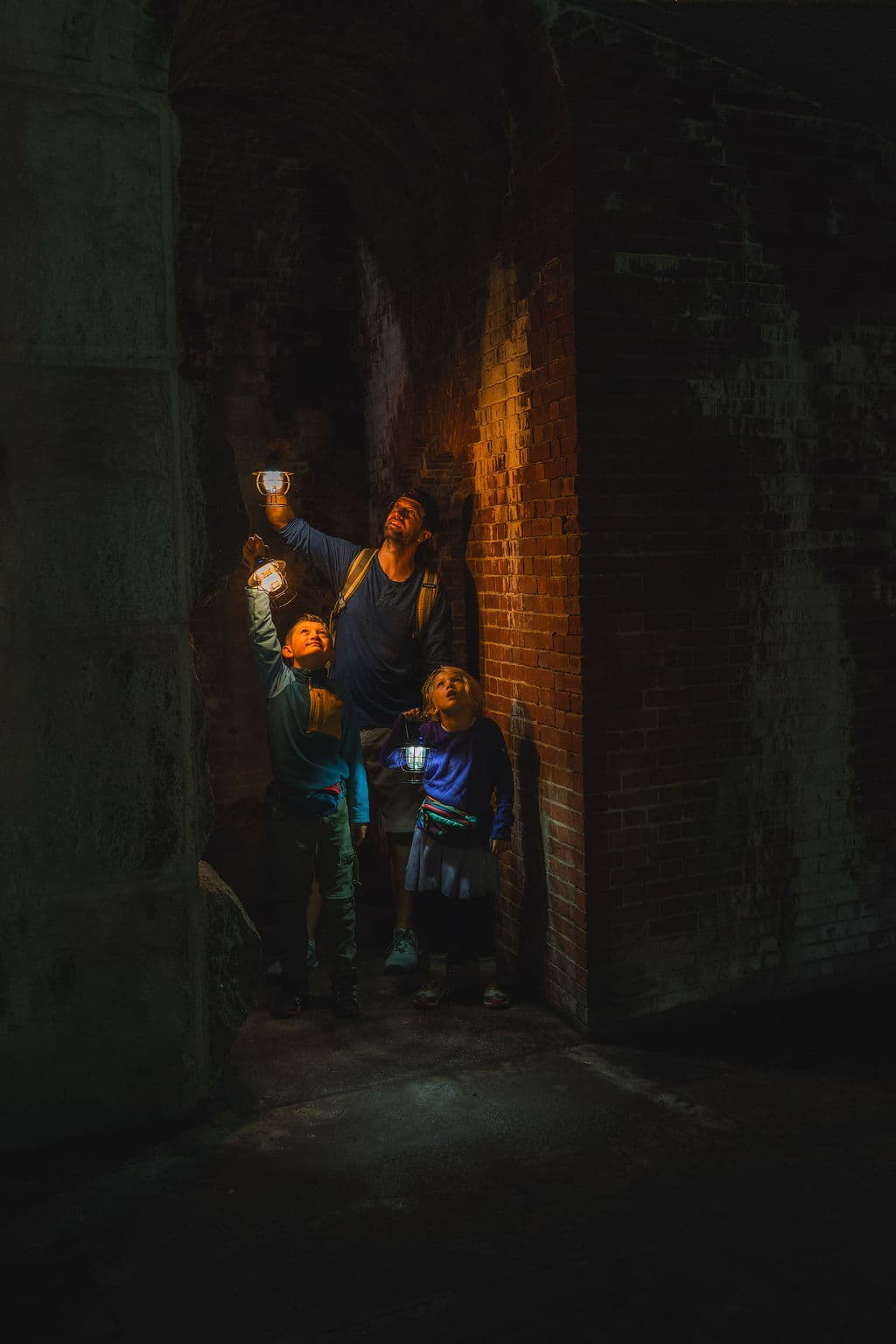 A man and two children explore a dimly lit space, holding up lanterns to illuminate their surroundings.