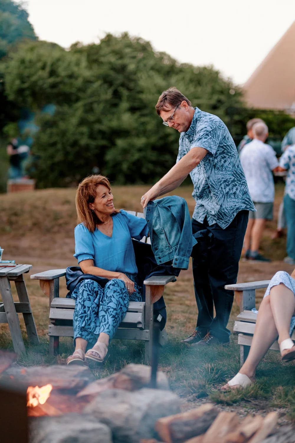 A man offers a jacket to a woman sitting outdoors by a fire pit, as others gather in the background.
