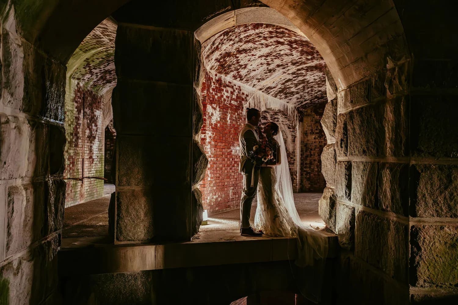 A couple embraces in a dimly lit, arching stone corridor adorned with rustic brick walls.