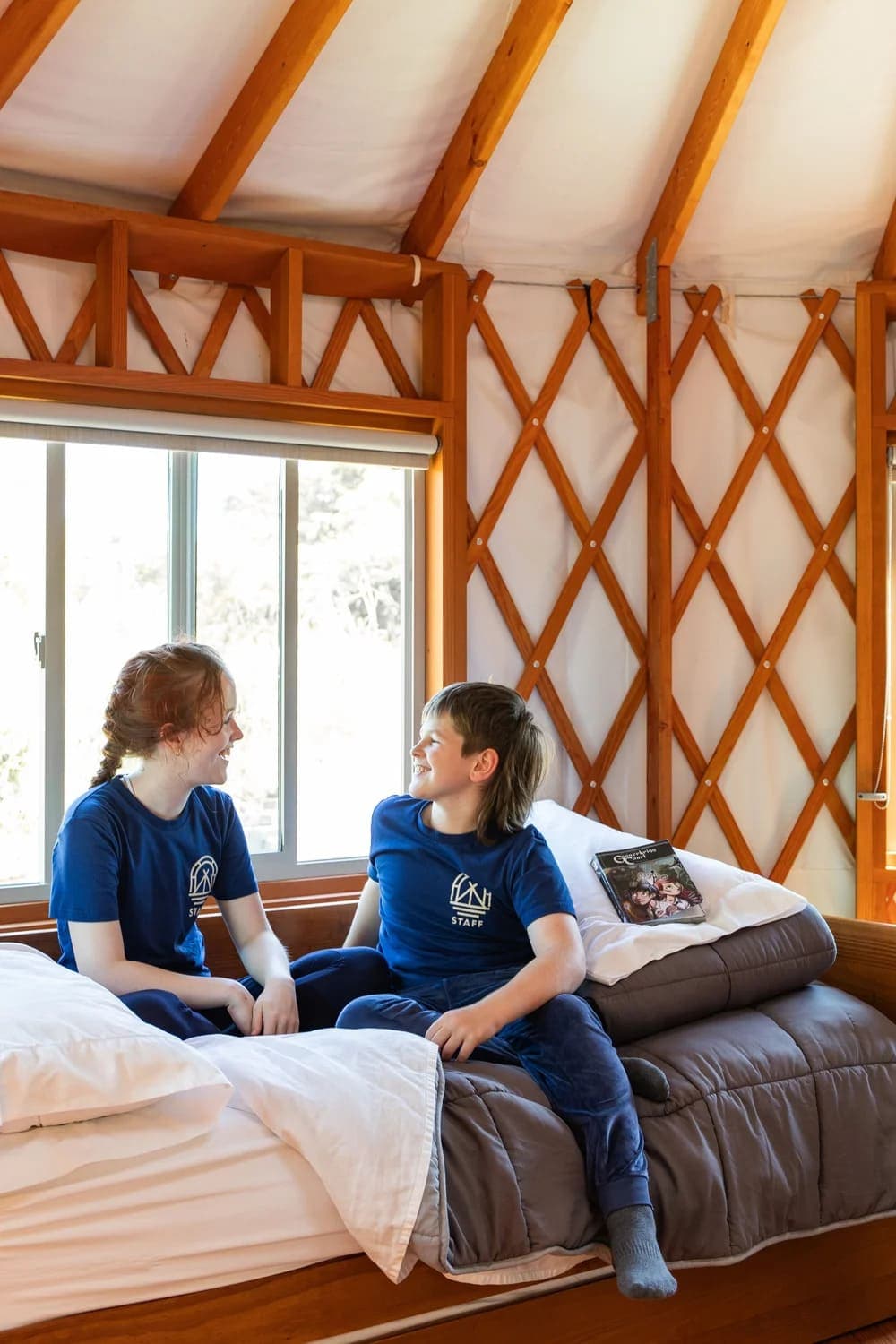 Two children in matching blue t-shirts sit on a bed inside a cabin, smiling at each other.