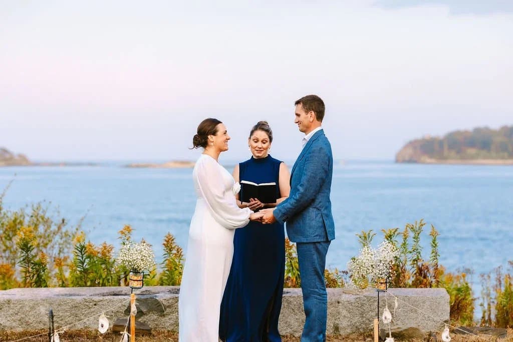A couple stands hand-in-hand during their wedding ceremony by the water, with a witness present.