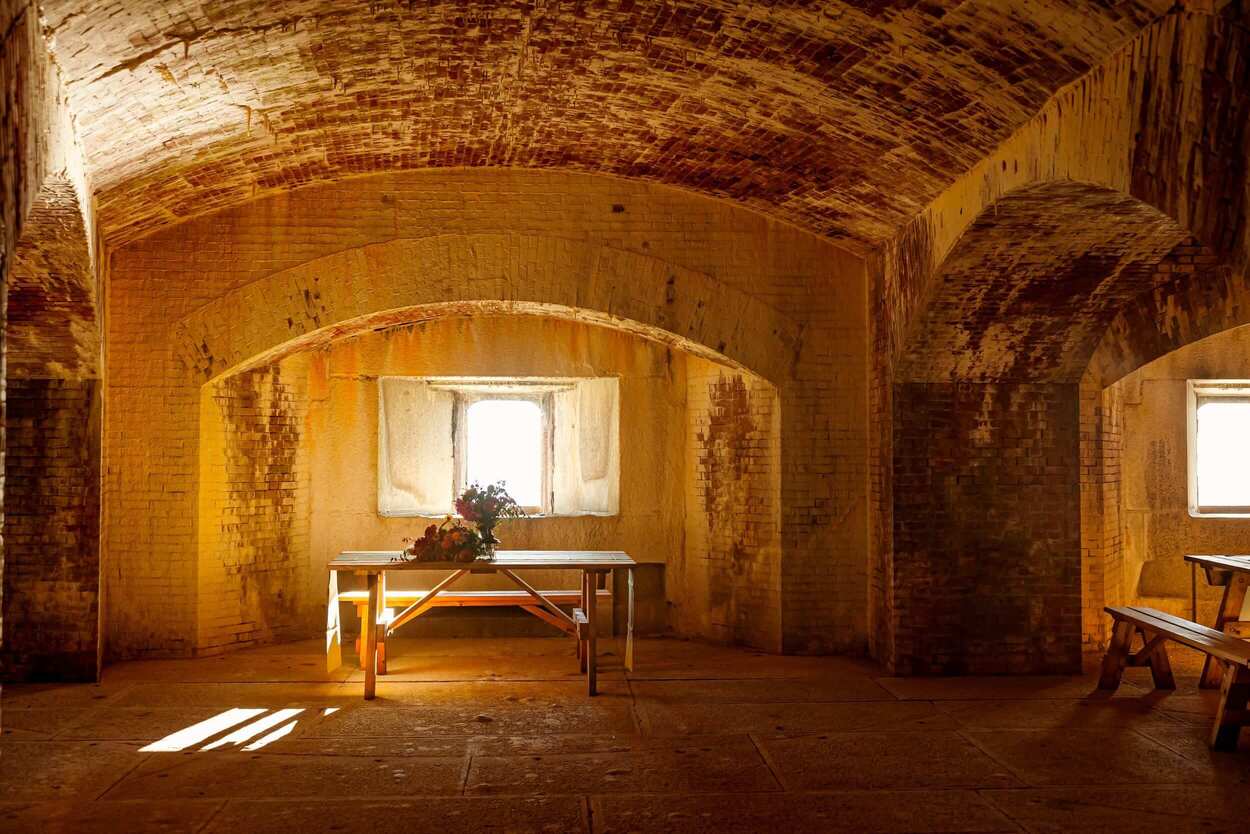 An empty, sunlit room with arched brick ceilings and a table adorned with flowers near a window.