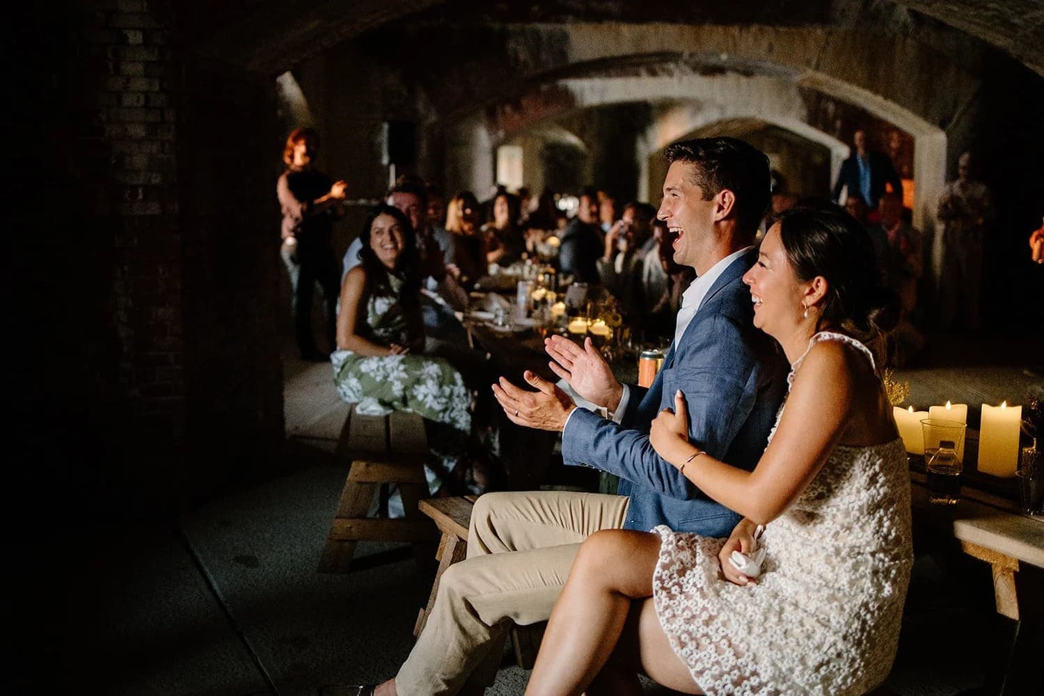 A man and woman laugh and applaud together while seated at a banquet in a candlelit venue, surrounded by guests.