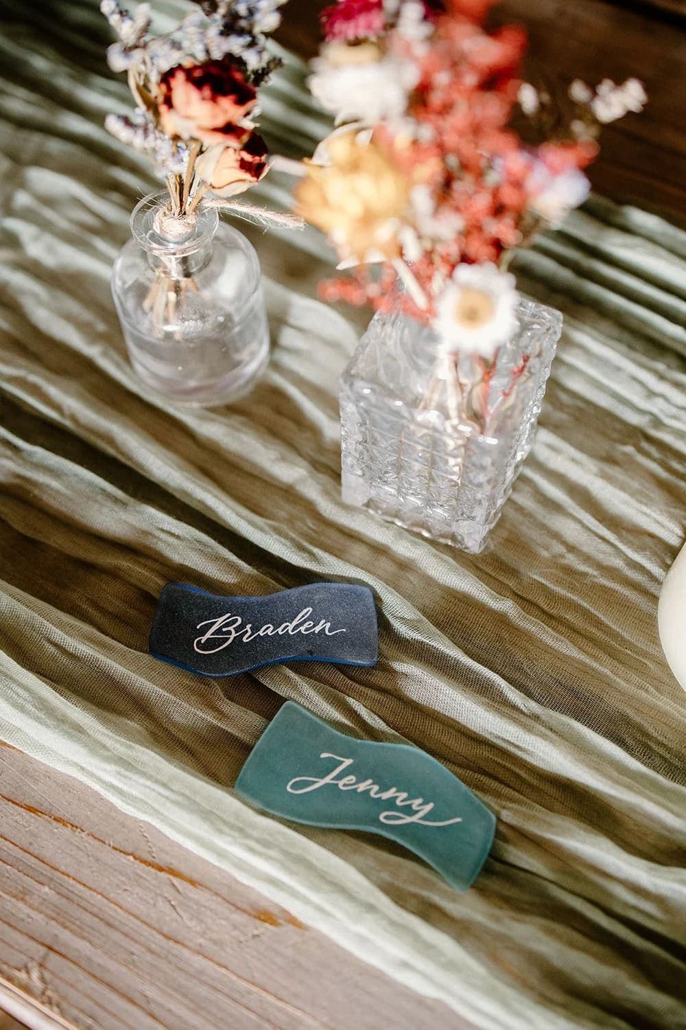 Two decorative name cards reading "Braden" and "Jenny" placed next to small vases of dried flowers on a textured table runner.