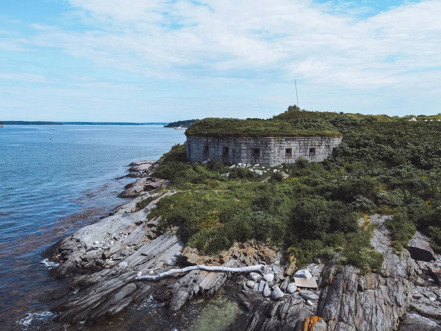 An old stone structure overgrown with vegetation sits along a rocky shoreline by the water.