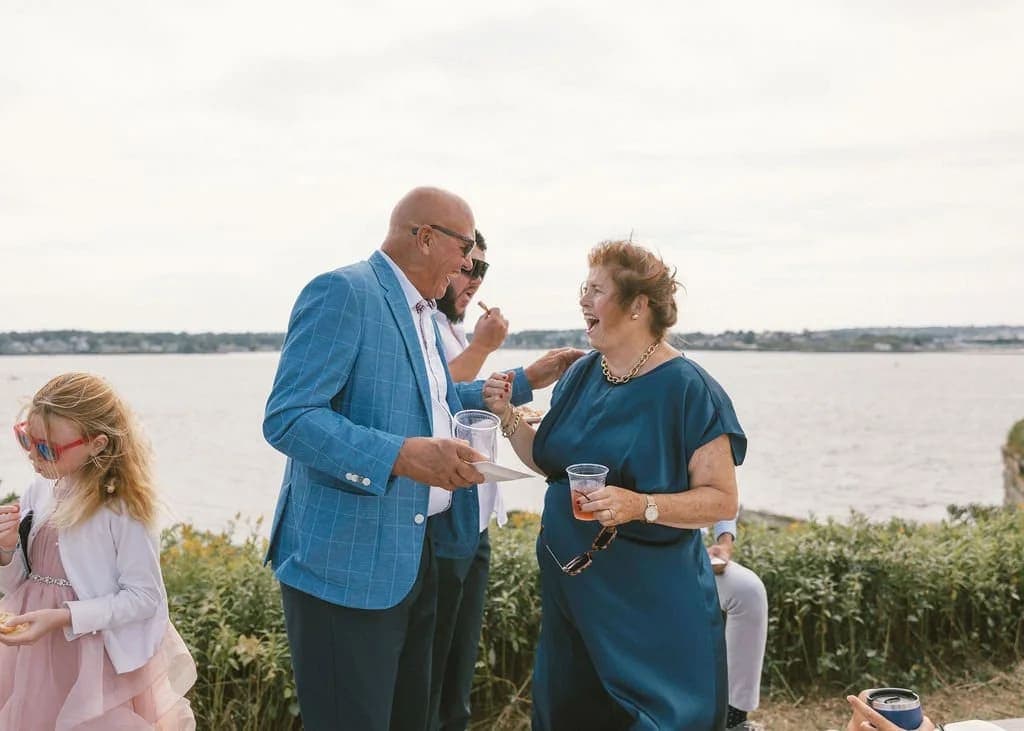 A man and a woman chat with drinks in hand, with a child nearby and a waterfront view in the background.