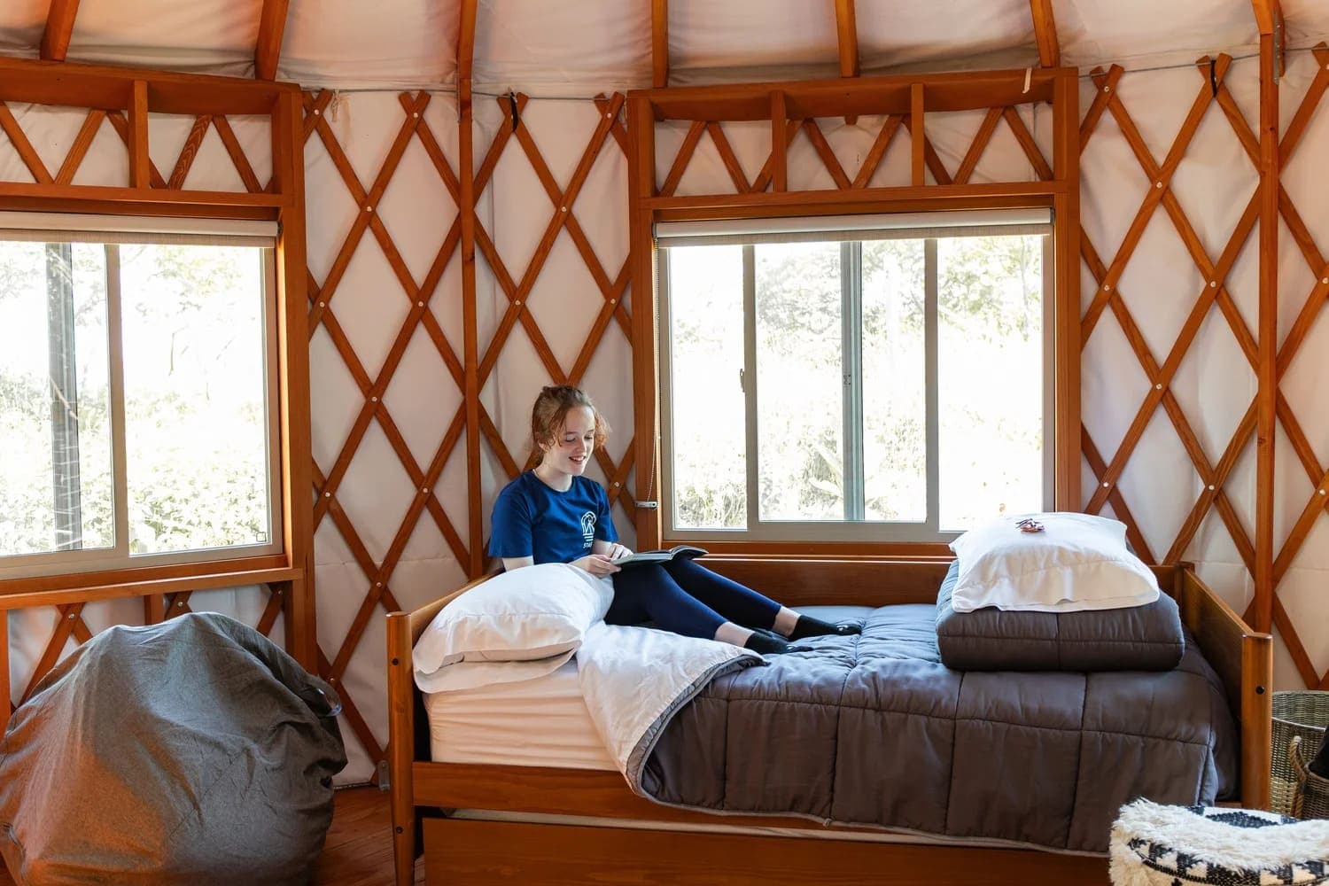 A child sits on a bed in a cozy yurt, reading a book by the window.