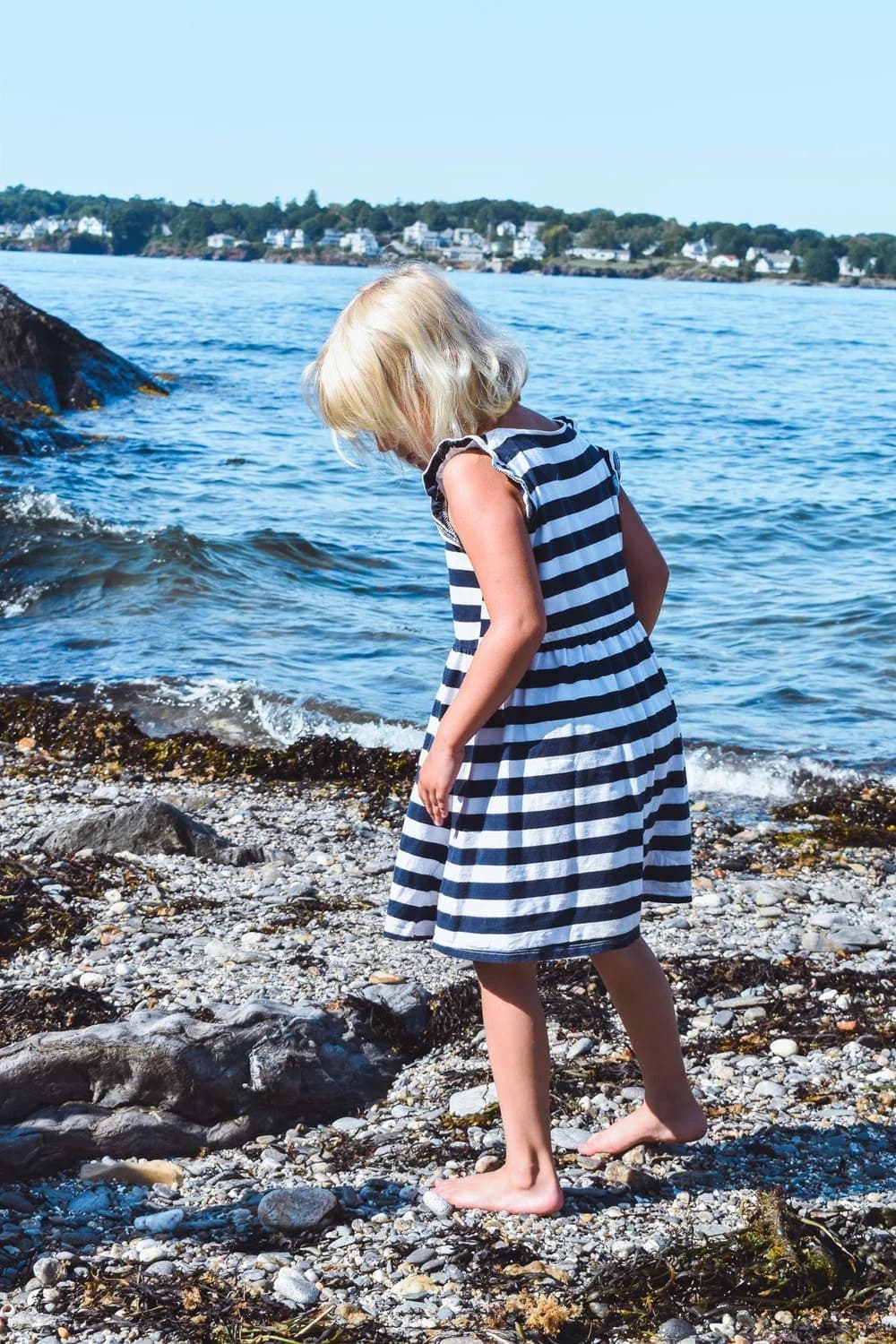 A young girl in a striped dress stands on a rocky beach, looking down at the water.