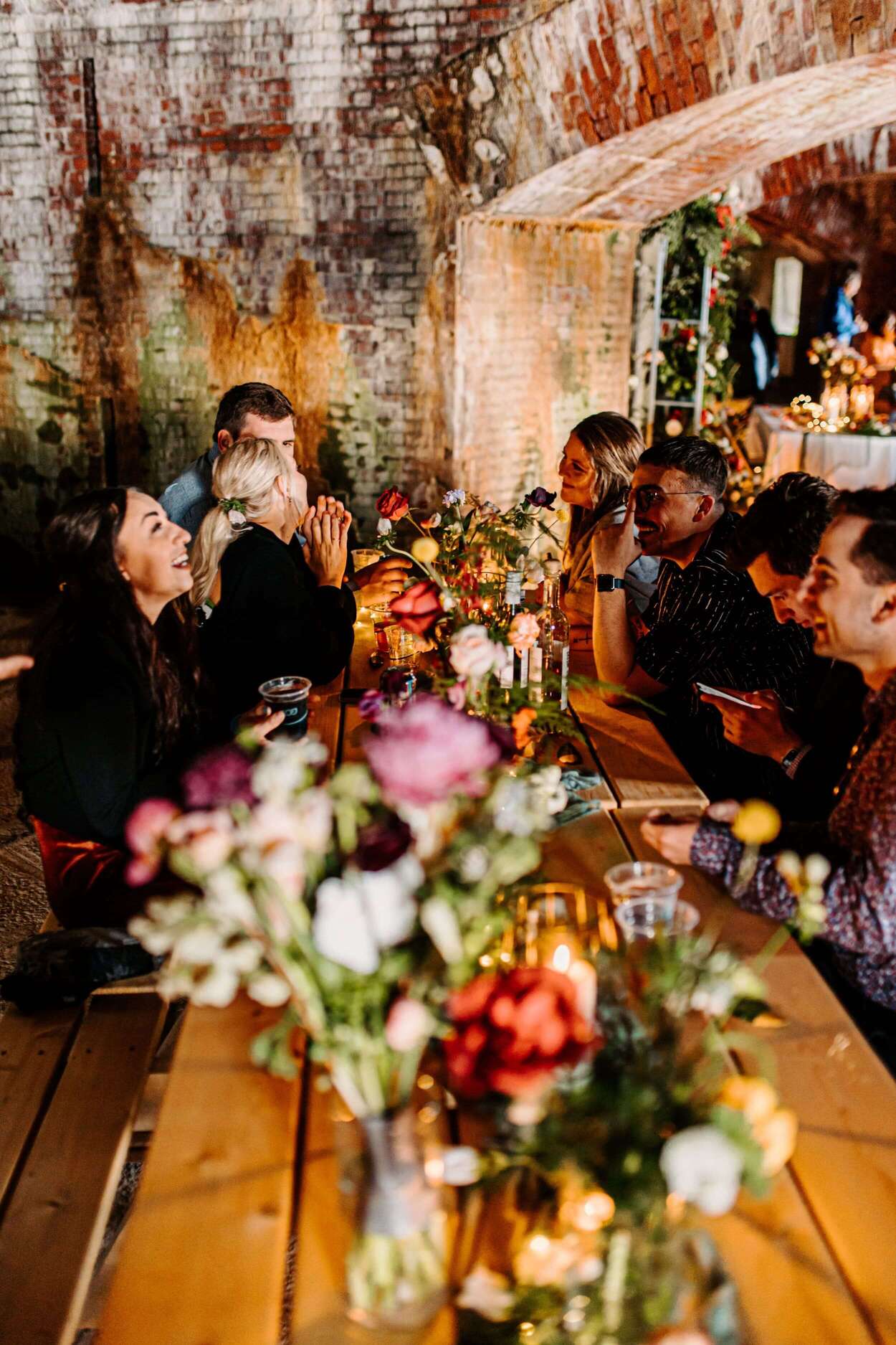 A group of people enjoy a lively conversation around a flower-adorned table in a rustic venue.
