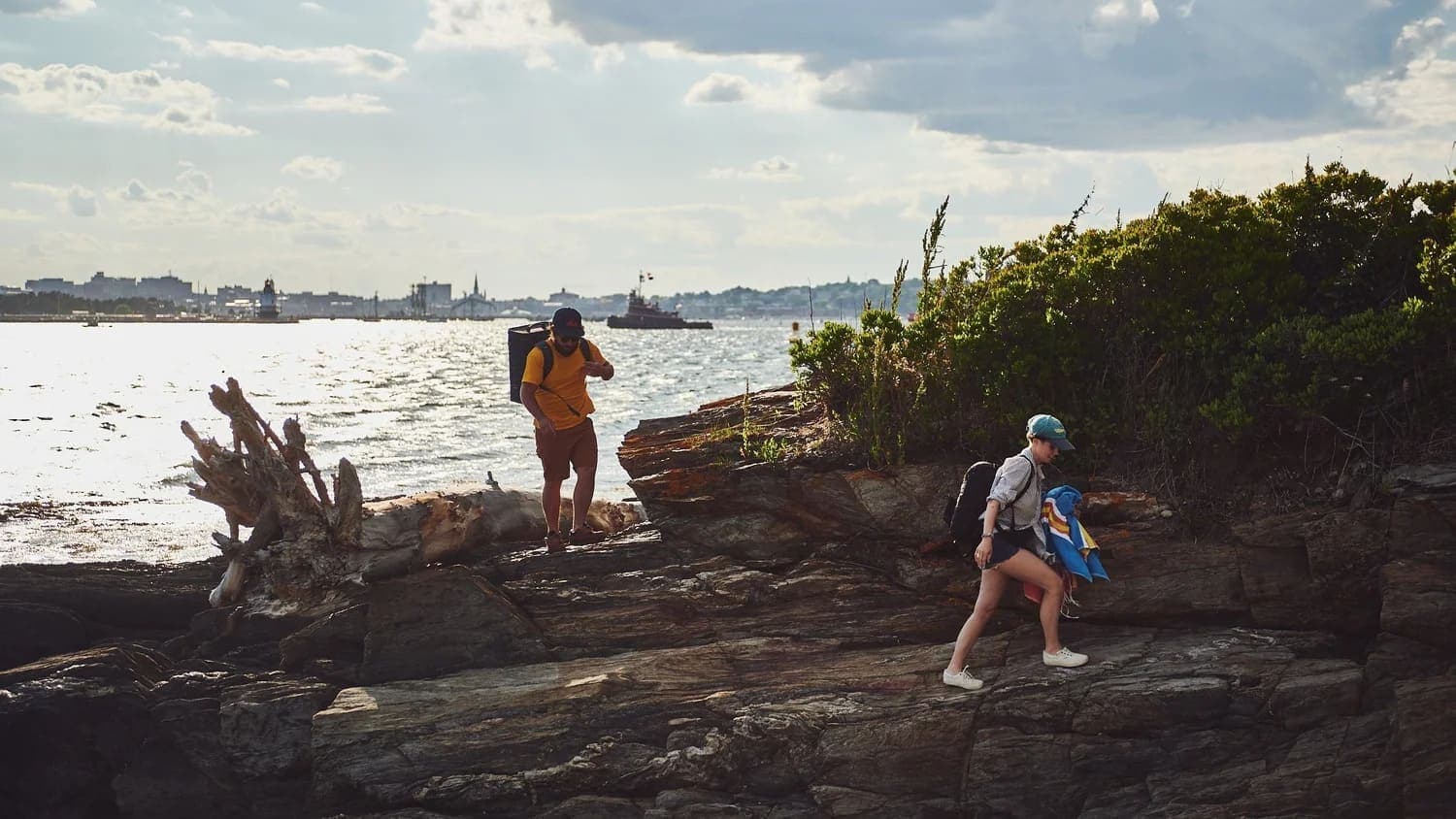 Two people hike along rocky terrain by the water, with a city skyline in the background.
