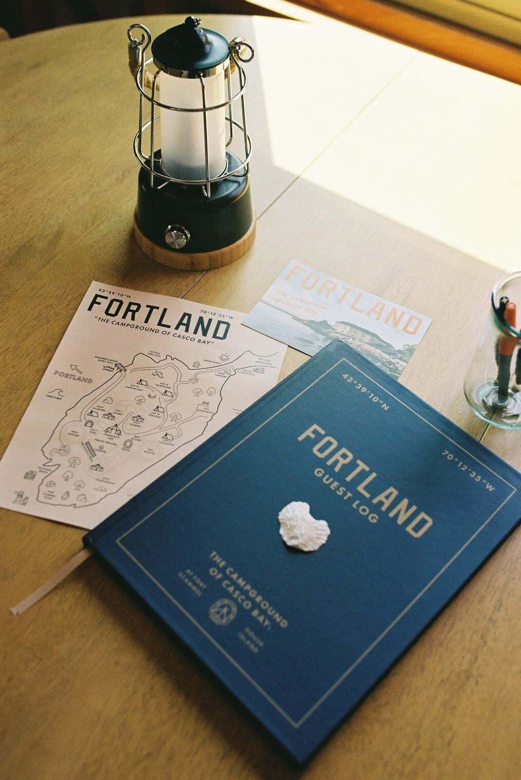 A vintage lantern and camping materials, including a map and guest log, are arranged on a wooden table.