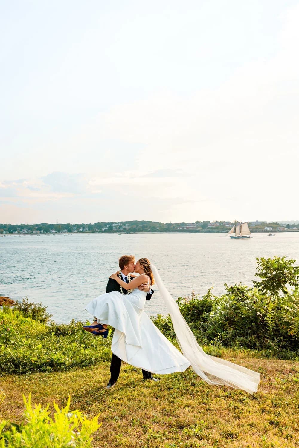 A newlywed couple sharing a kiss, with the bride being lifted in a scenic waterfront setting.