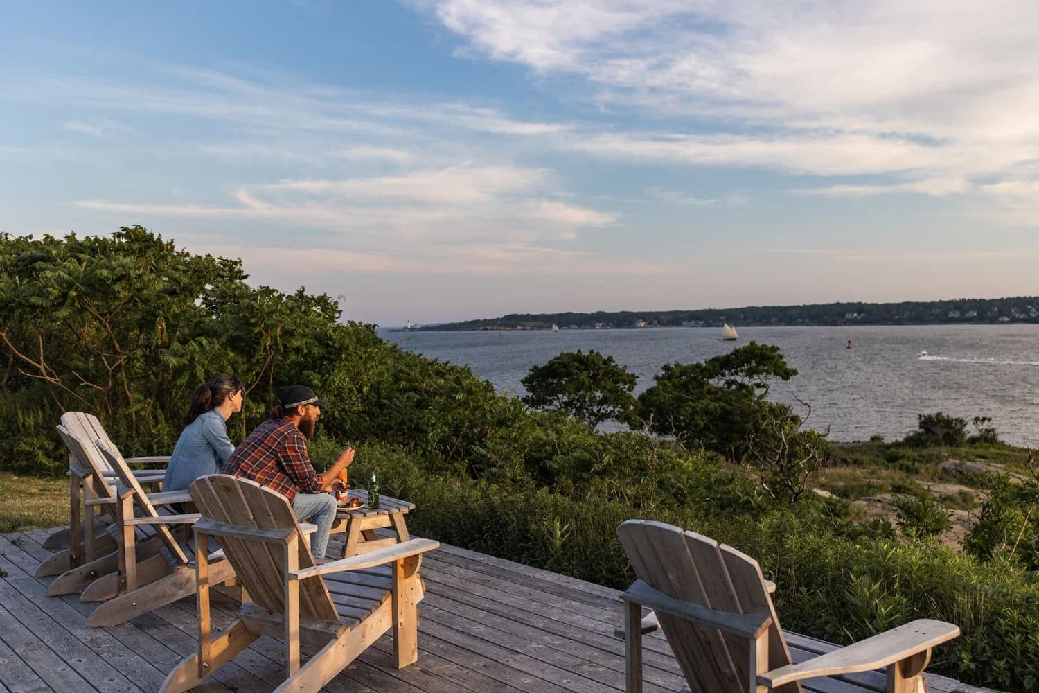 Two people sit in wooden chairs on a deck overlooking a calm bay at sunset.