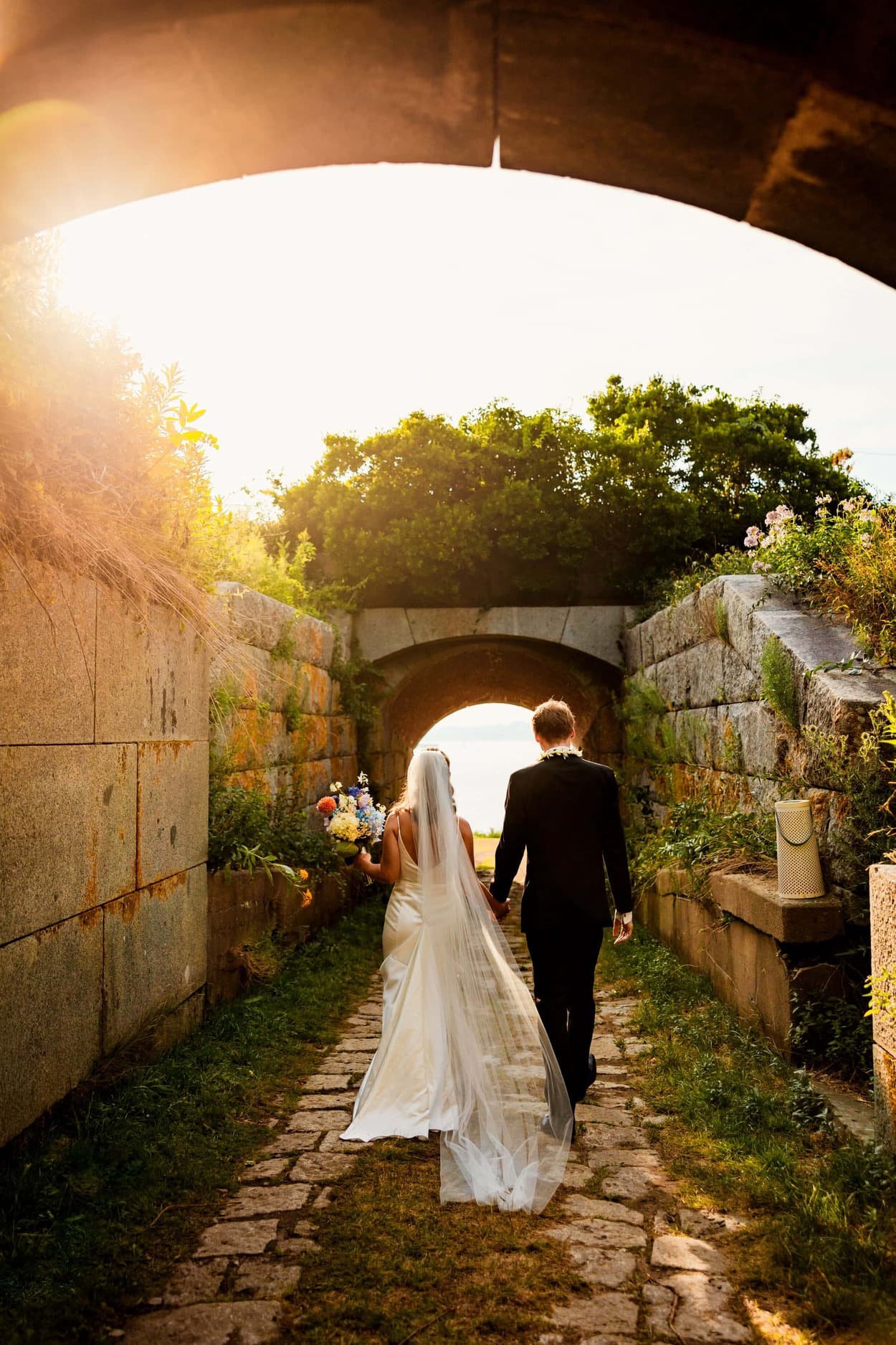 A couple walks hand in hand down a sunlit stone path beneath a lush archway.