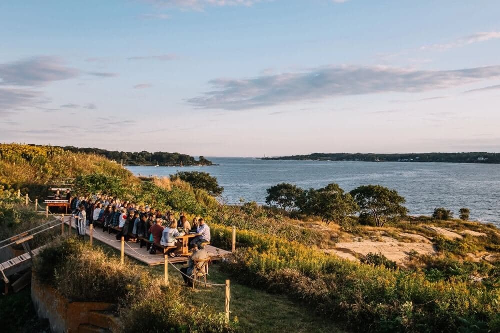 A large group of people dining outdoors by the water at sunset.