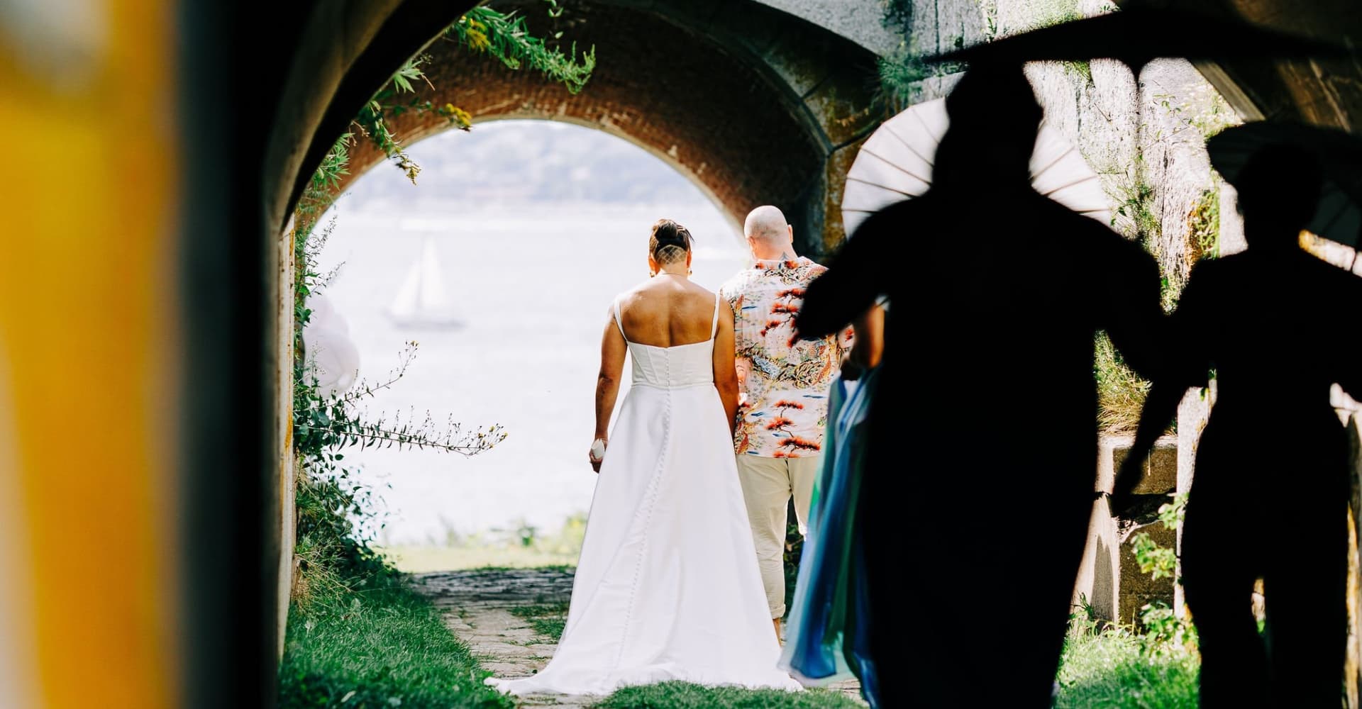 A bride in a white dress walks toward a groom through an arched pathway, with silhouettes of guests in the foreground.