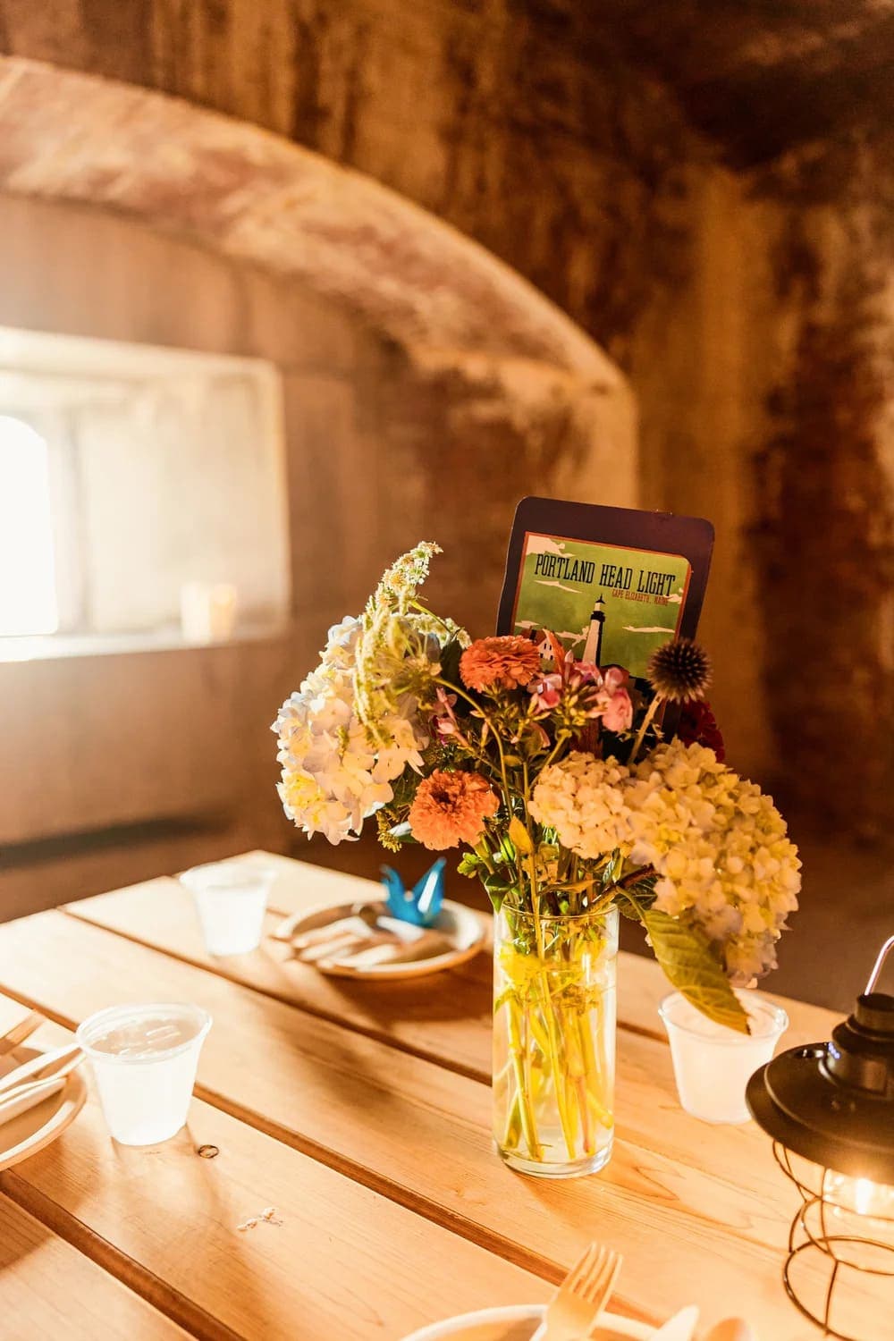 A clear vase with colorful flowers and a sign reading "Portland Head Light" on a wooden table in a softly lit room.