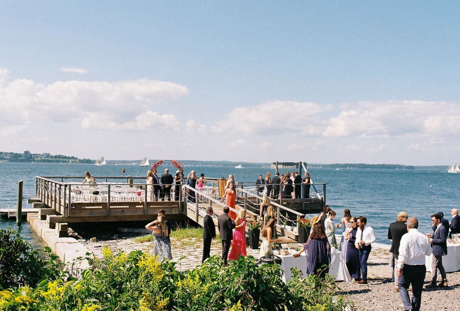 A waterfront wedding reception with guests mingling near a dock under a clear blue sky.