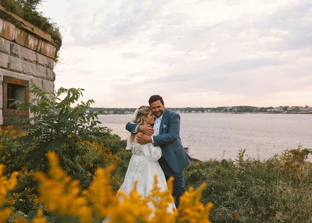 A couple embraces by a body of water, surrounded by flowers and a historic stone structure.
