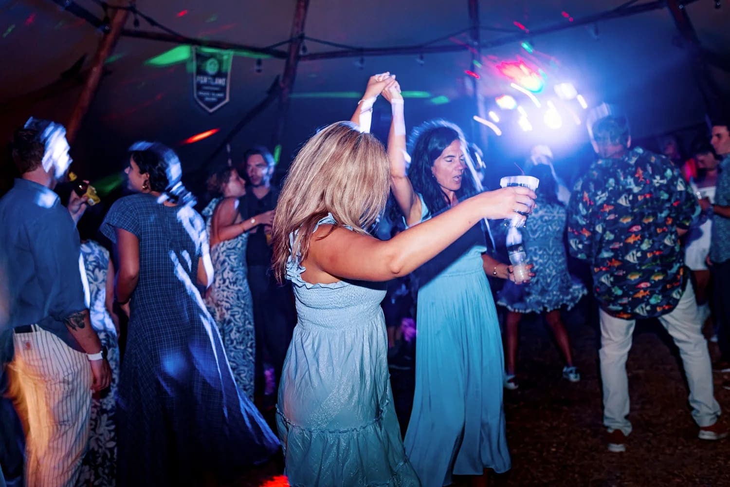 A lively group of people dances under colorful lights in a festive tent.