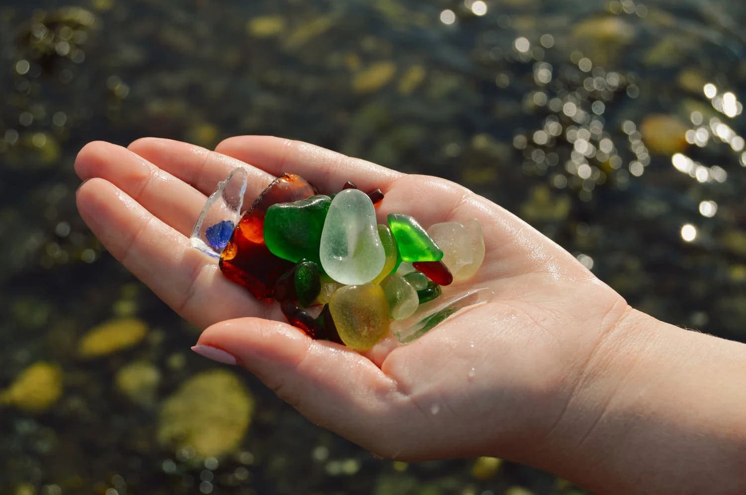 A hand holds a collection of colorful sea glass pieces above a shimmering body of water.