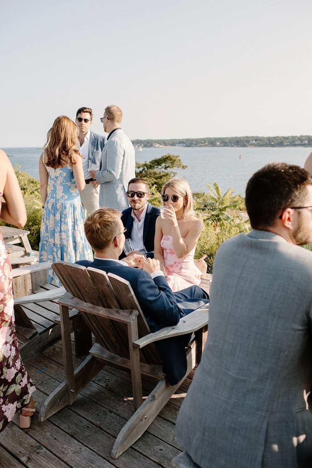 A group of people socialize on a deck overlooking a body of water on a sunny day.