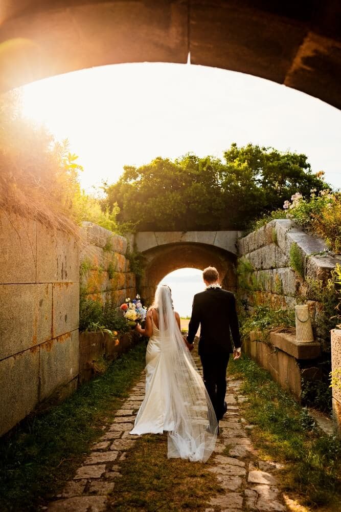A bride and groom walk hand in hand through a sunlit stone pathway beneath an archway.