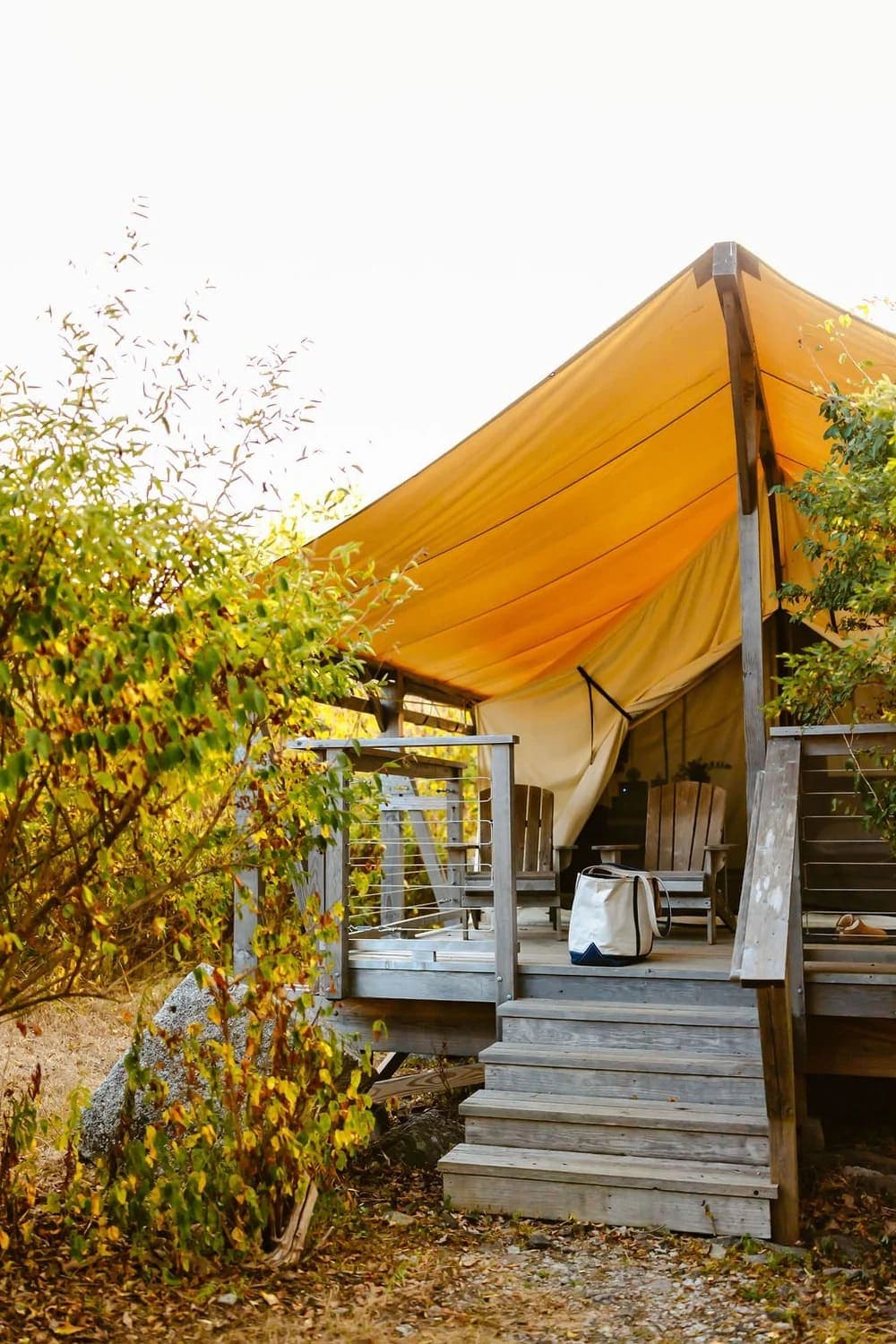 A cozy glamping tent with a yellow canopy and wooden deck, surrounded by autumn foliage.