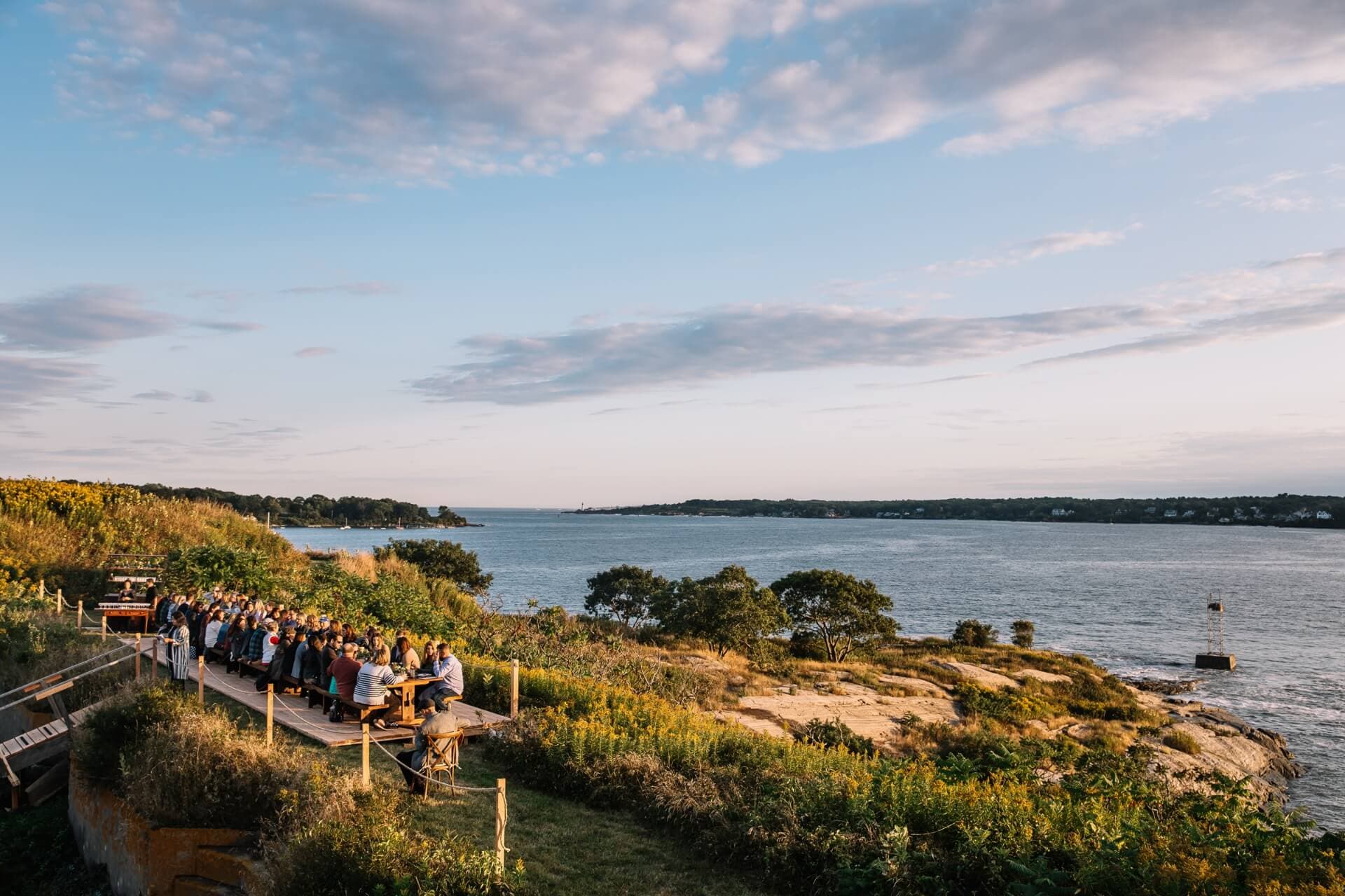 A group of people sits at a dining table overlooking a serene coastline at sunset.