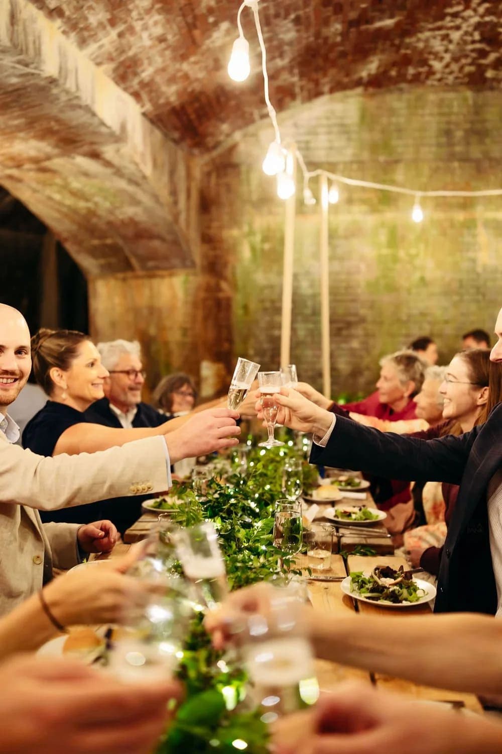 A lively group toasts at a long dining table adorned with greenery in a rustic, warmly lit setting.