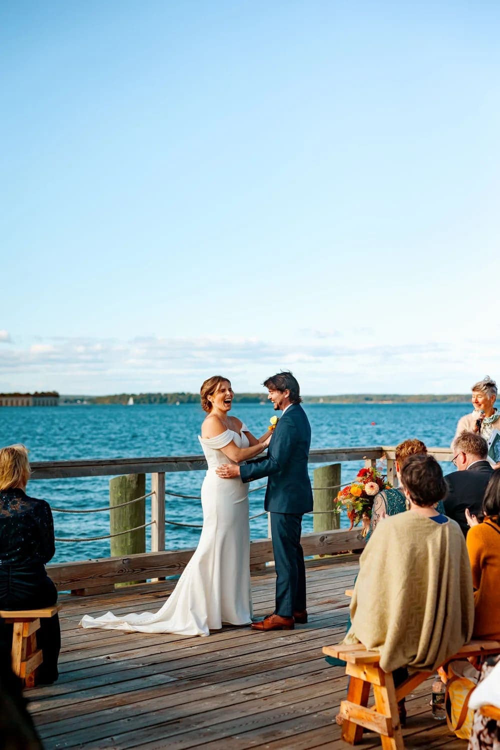 A bride and groom exchange vows on a waterfront deck surrounded by guests.