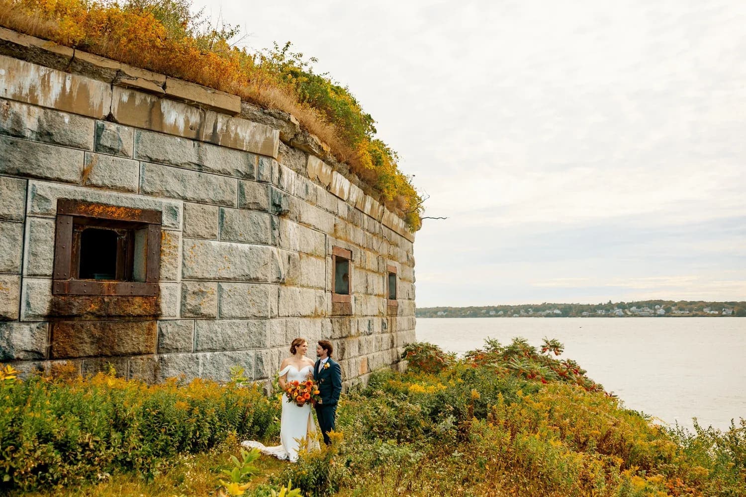 A couple in wedding attire stands amidst overgrown greenery near a stone structure by the water.