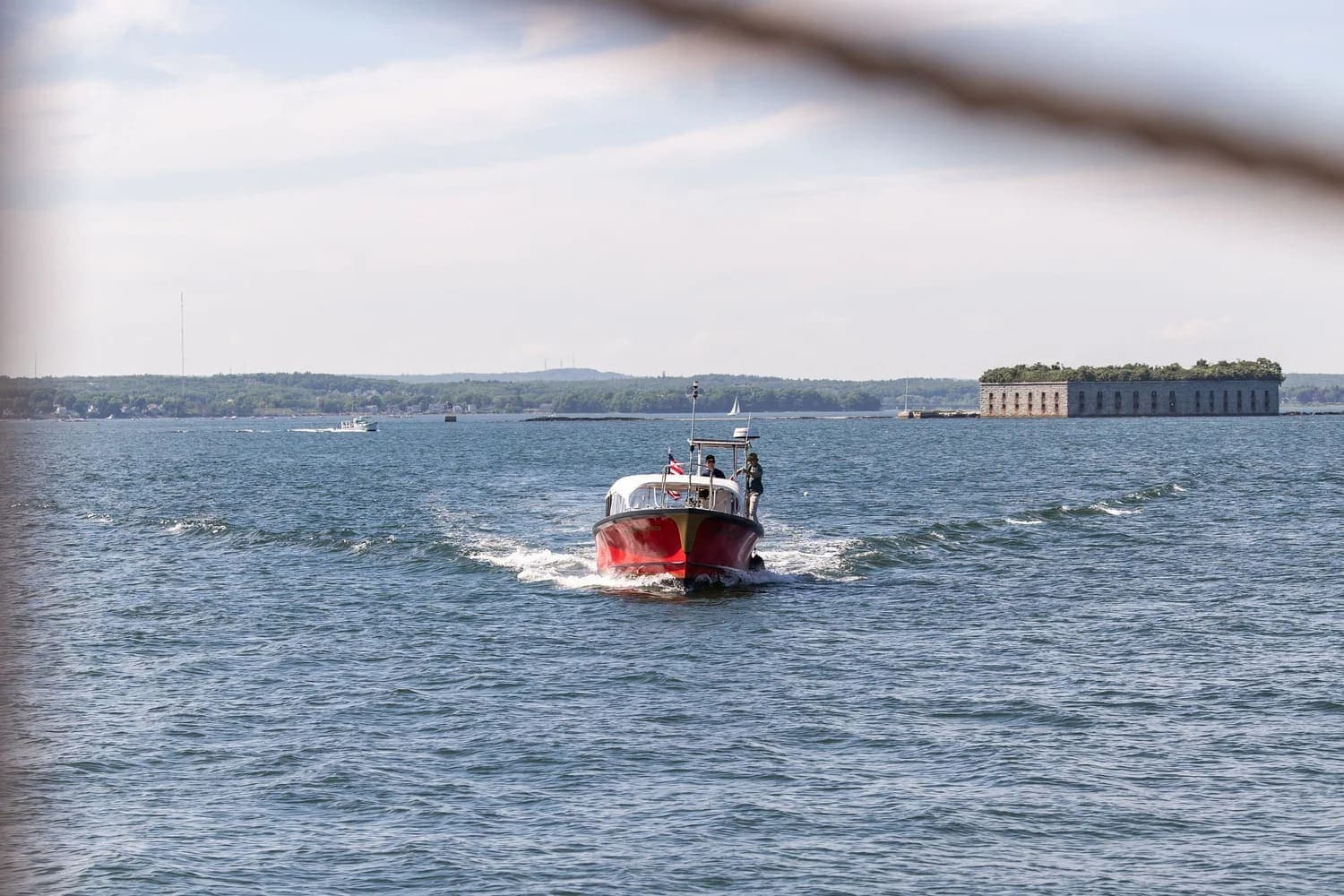 A small red boat sails through calm waters near a historic fort.